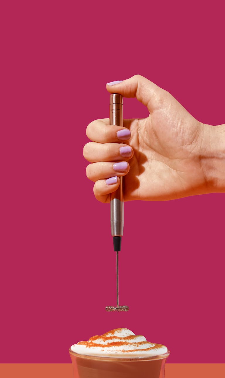 A hand frothing milk in coffee with a vibrant pink backdrop, showcasing a modern lifestyle.