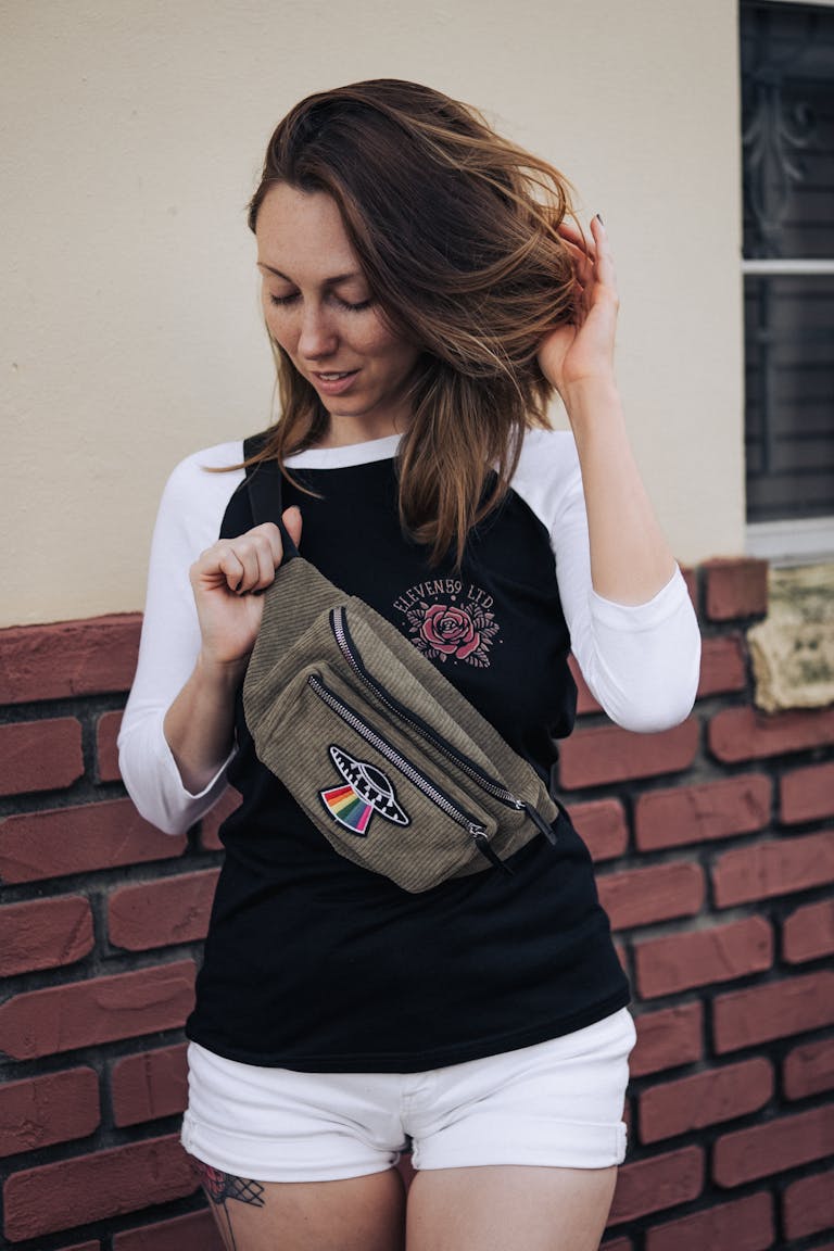 Caucasian woman posing stylishly in casual outfit with a belt bag against a brick wall.