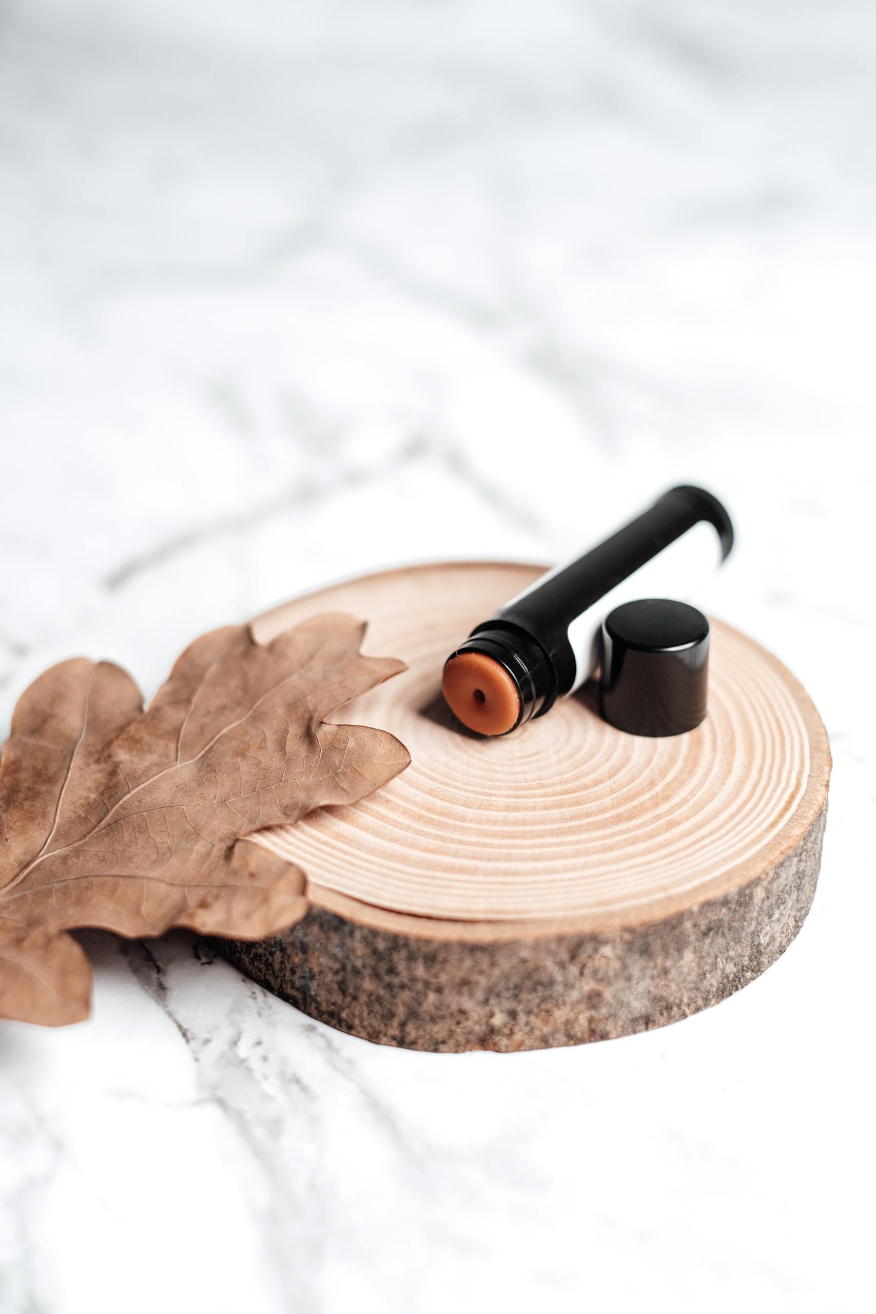 Close-up of lip balm on a wooden slice against a marble background, featuring a dried leaf.