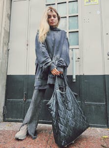 Fashionable woman standing on a city sidewalk holding a quilted bag in an edgy outfit.