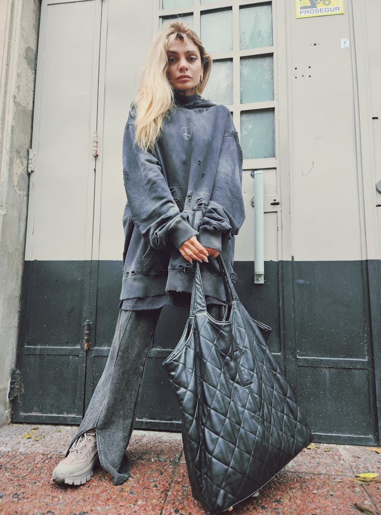 Fashionable woman standing on a city sidewalk holding a quilted bag in an edgy outfit.