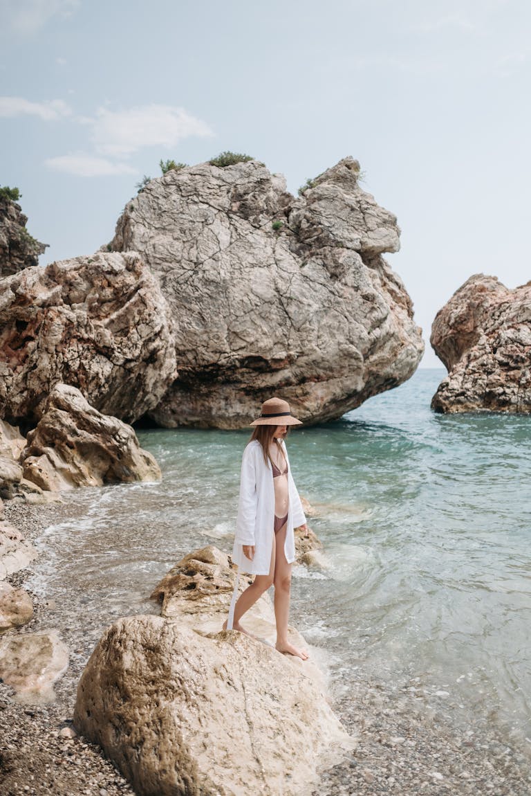 Woman in a hat and swimsuit walking by large rocks on a serene beach.