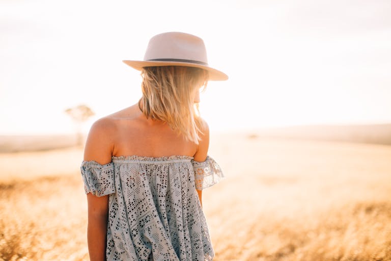 A blonde woman stands in a sunlit field wearing a stylish dress and hat, embodying summer fashion.