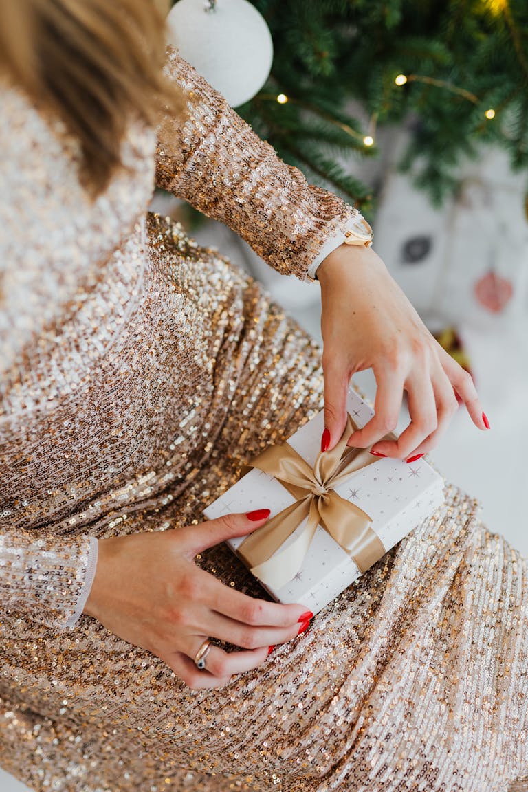 An elegant woman opens a wrapped gift, showcasing holiday spirit in a sparkling dress.
