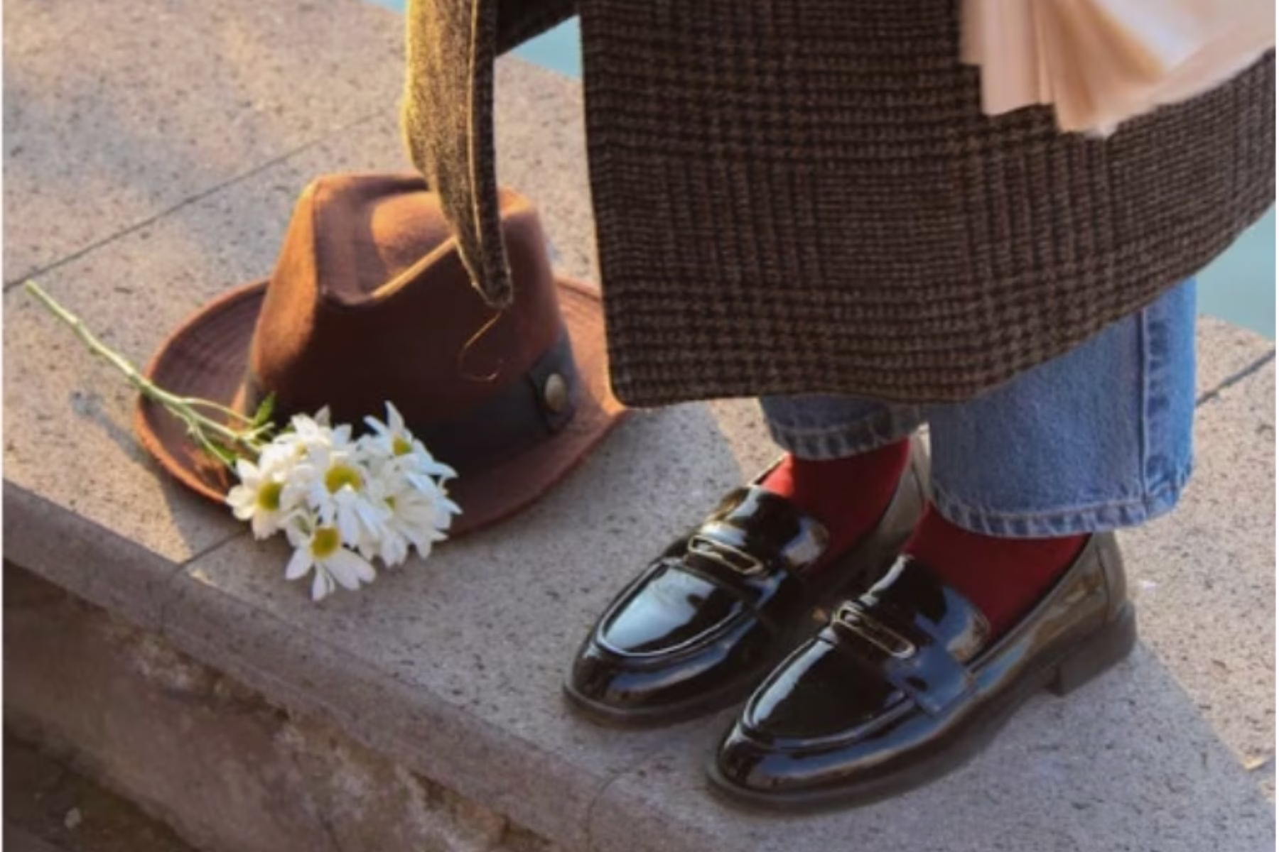 woman in trench wearing loafers with a hat next to her